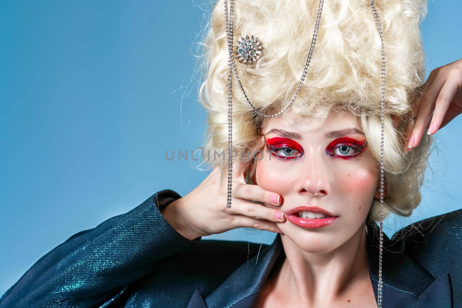 Portrait of funny princess with vintage wig and red makeup making faces. Woman wearing elegant jacket on blue background. Royal party in blue studio.