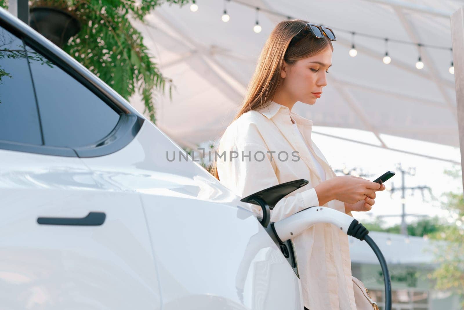 Sustainable urban commute with young woman pay electricity for EV electric car recharging at outdoor cafe in springtime garden, green city sustainability and environmental friendly EV car. Expedient
