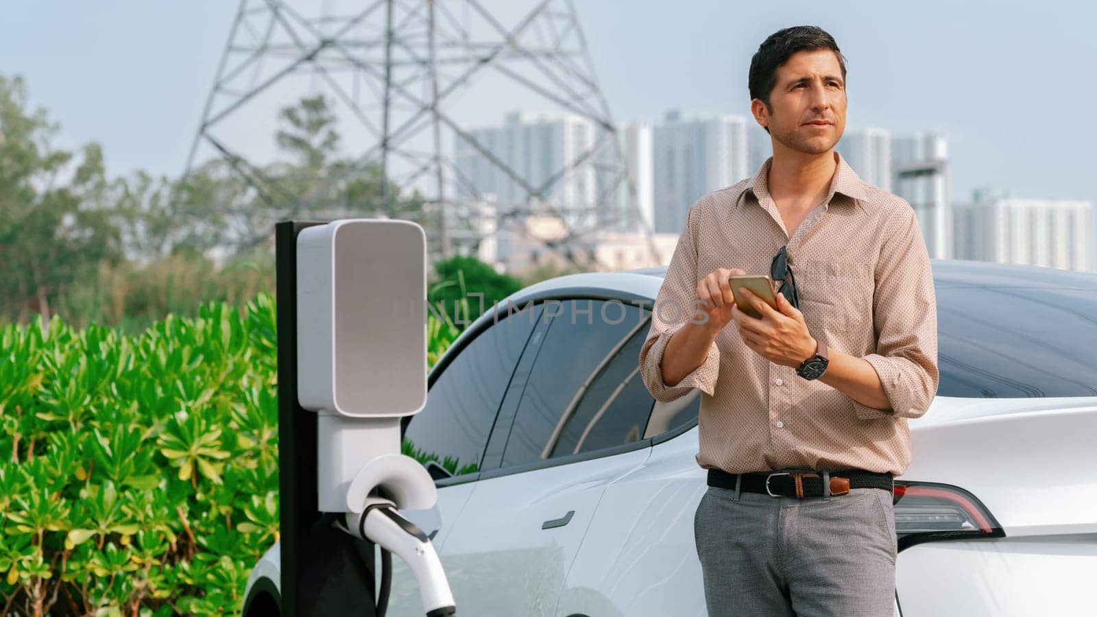 Man pay for electricity with smartphone while recharge EV car battery at charging station connected to power grid tower electrical as electrical industry for eco friendly car utilization.Expedient