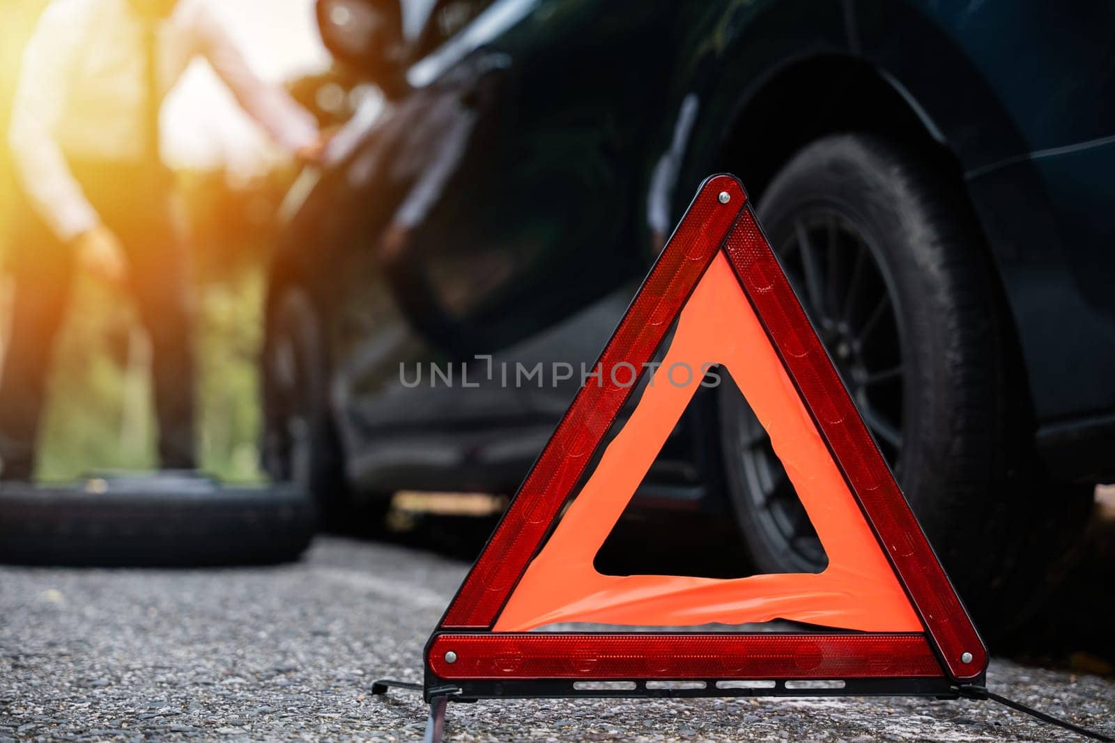 Businessman stranded on the road with a broken car and a warning triangle sign. Repairing the car wheel while waiting for roadside assistance. Concept of transportation problems and emergency support.