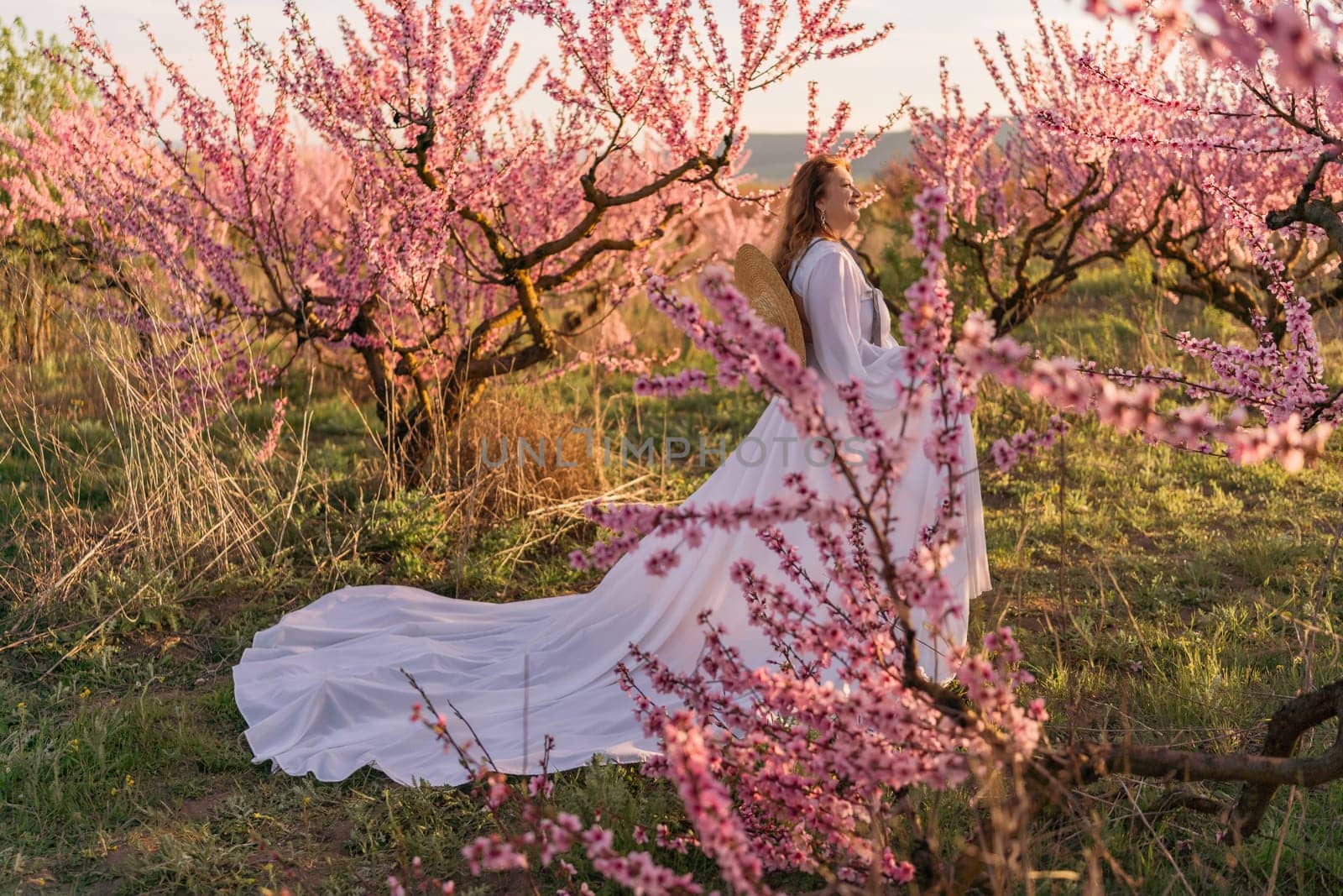 Woman blooming peach orchard. Against the backdrop of a picturesque peach orchard, a woman in a long white dress and hat enjoys a peaceful walk in the park, surrounded by the beauty of nature