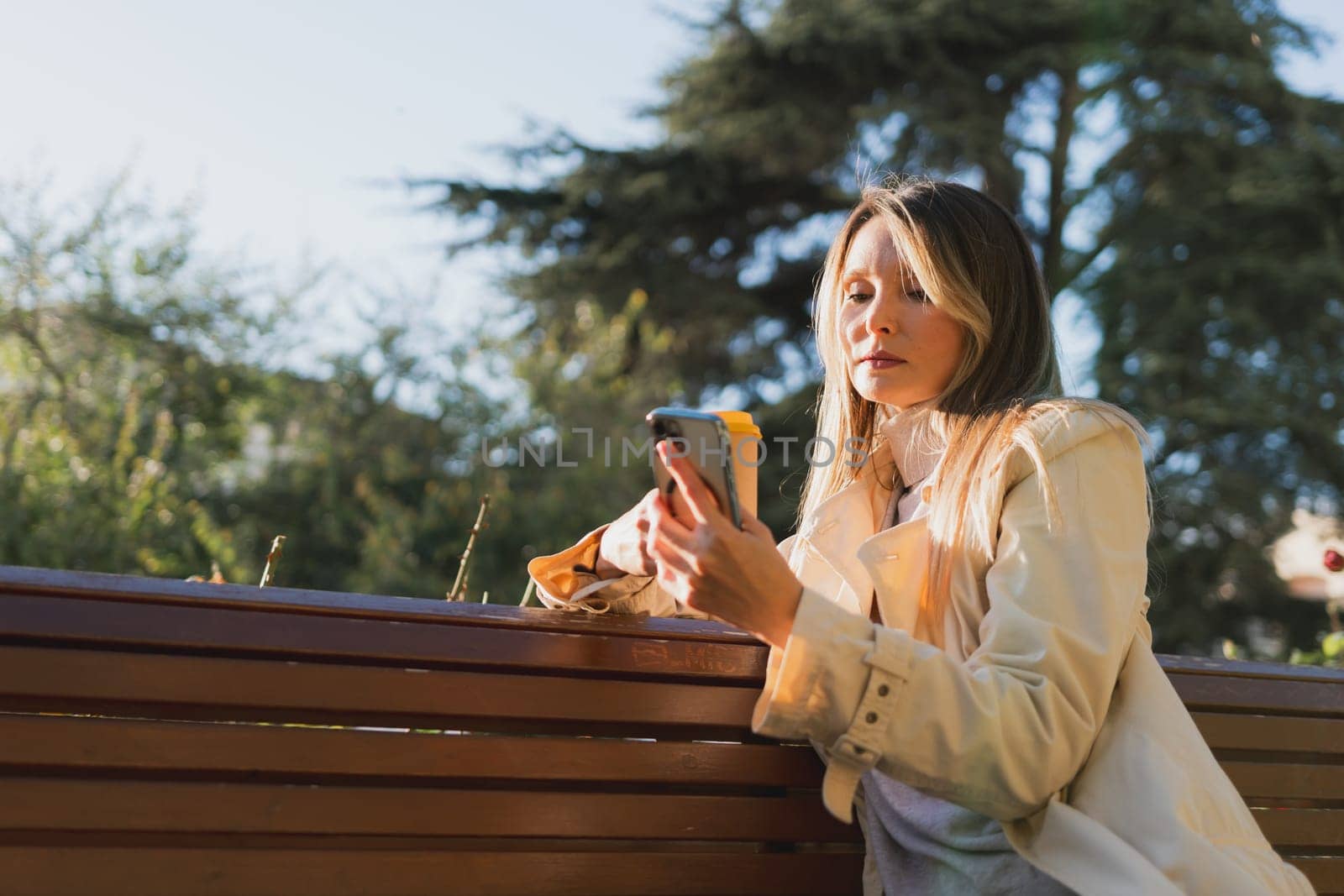 Woman drinks from cup on wooden bench. She is wearing a white shirt enjoying her beverage. The bench is located in a park setting, with trees in the background