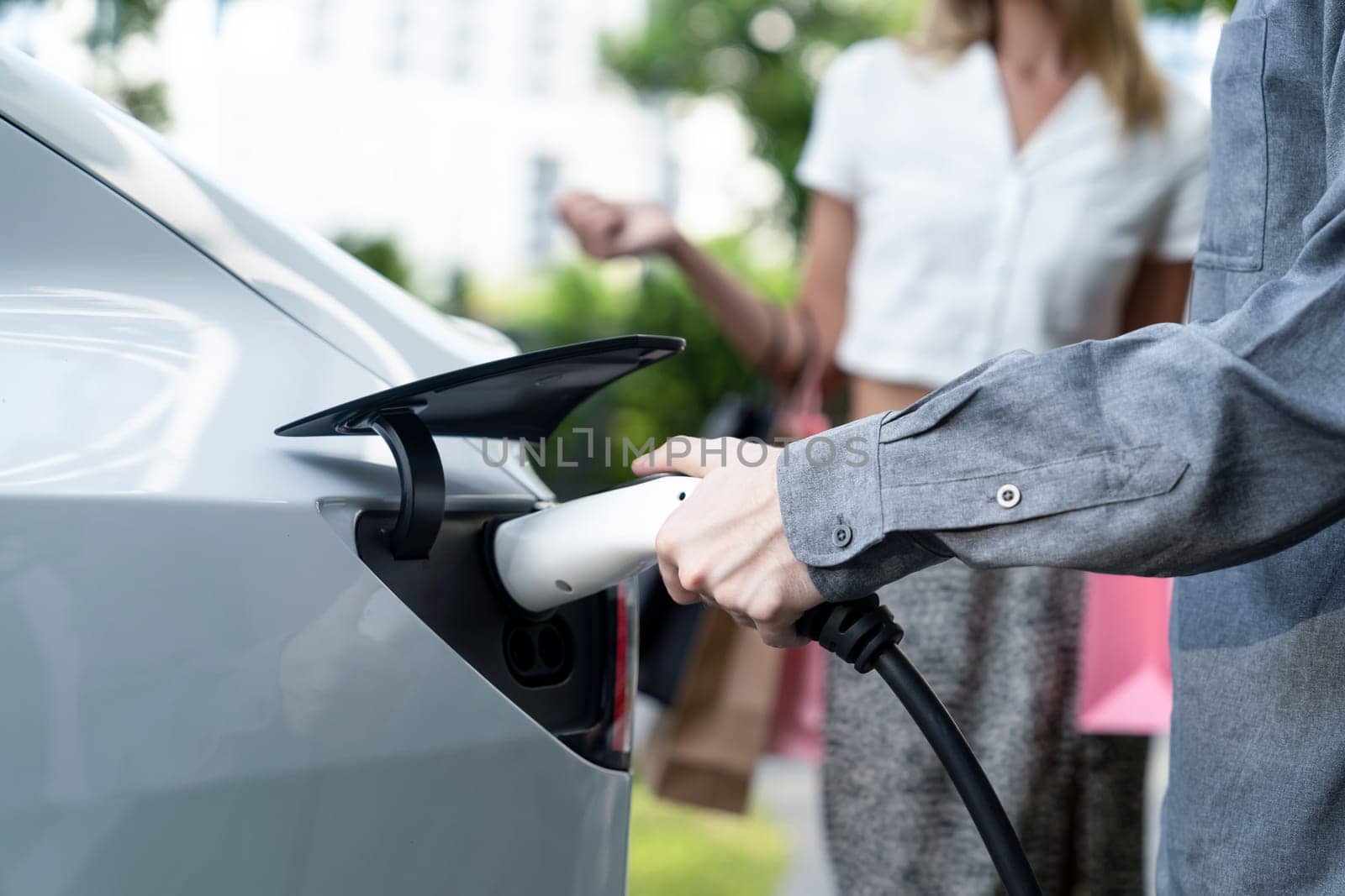 Young couple travel with EV electric car charging in green sustainable city outdoor garden in summer shows urban sustainability lifestyle by green clean rechargeable energy of electric vehicle innards