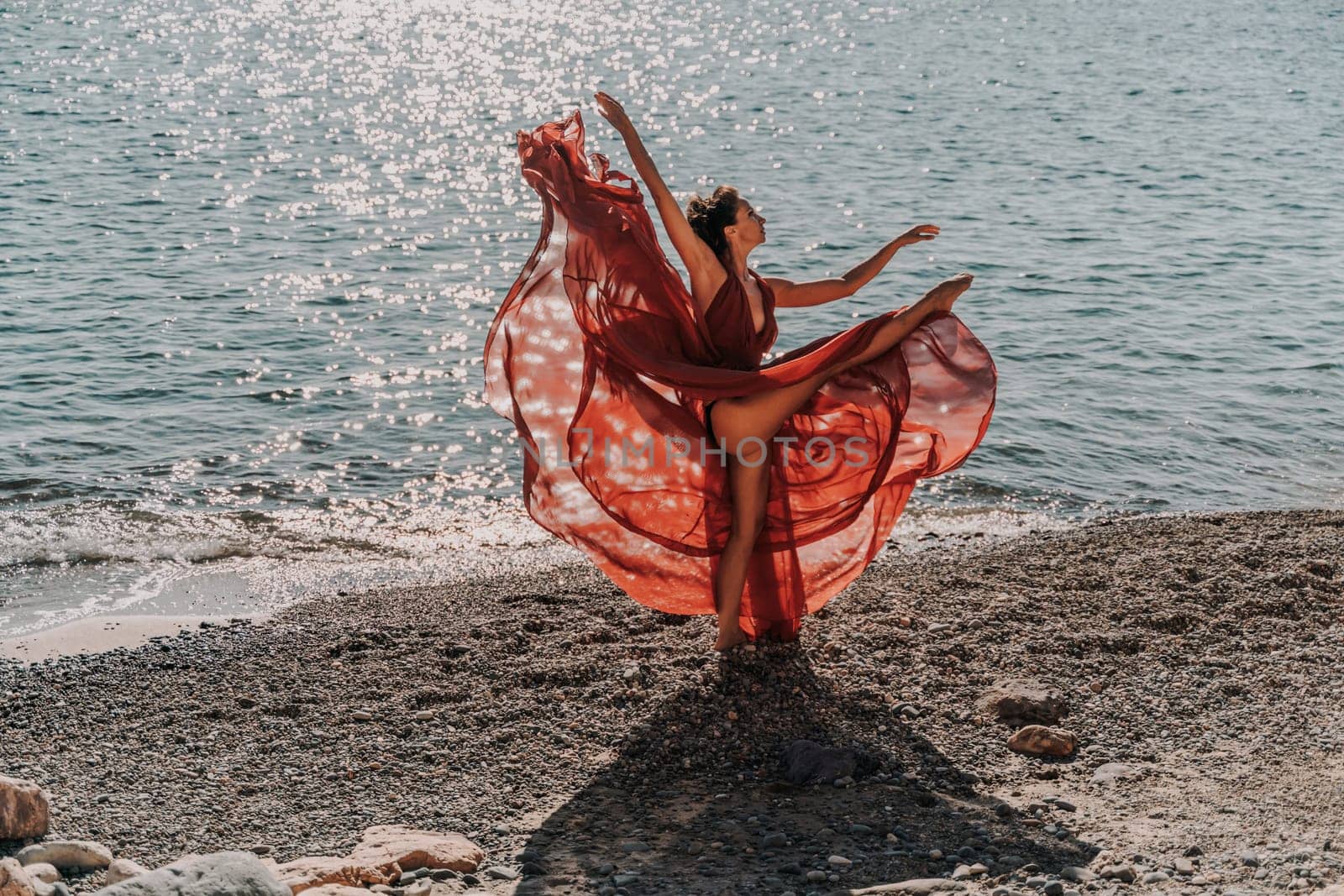 Woman red dress sea. Female dancer in a long red dress posing on a beach with rocks on sunny day. Girl on the nature on blue sky background. by Matiunina