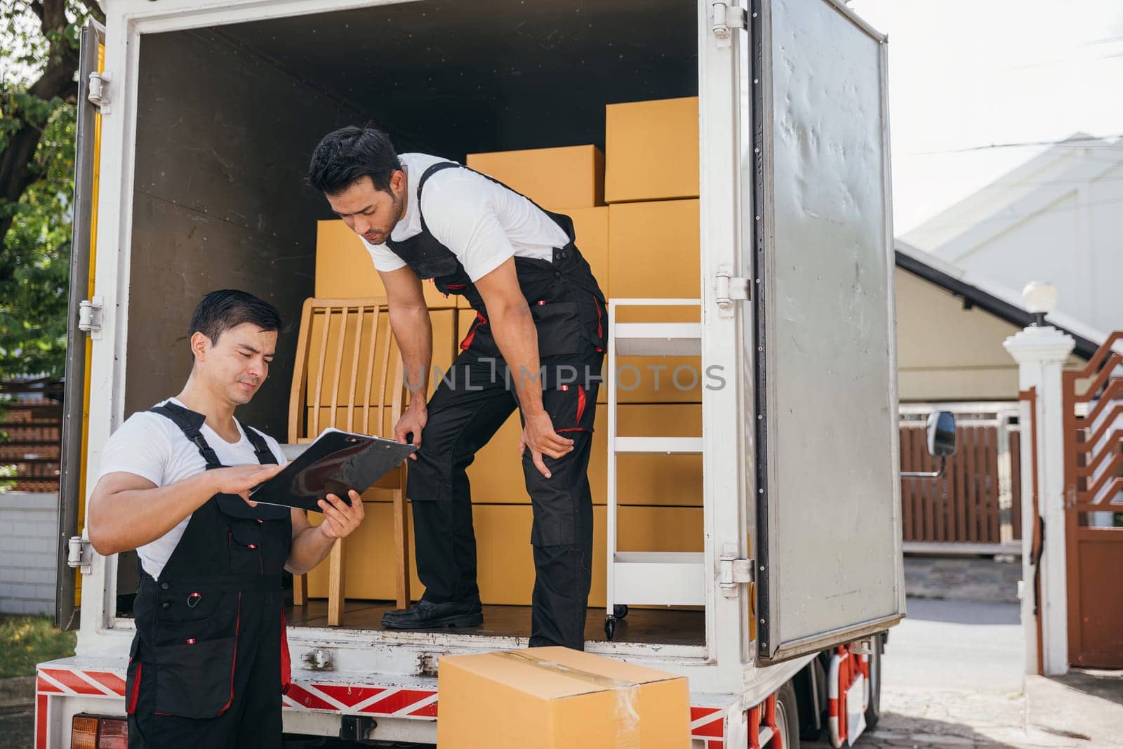 Mover workers unload boxes inspecting shipment with a clipboard near the truck. Professional delivery team ensures seamless relocation and efficient service. Moving day concept by Sorapop