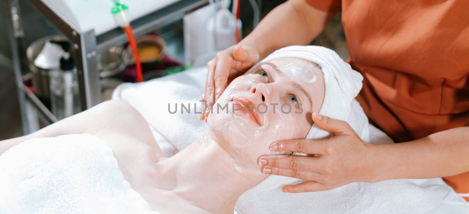 Portrait of beautiful caucasian woman having facial massage with homemade facial mask while lies on spa bed surrounded by beauty electrical equipment and peaceful nature environment. Tranquility.