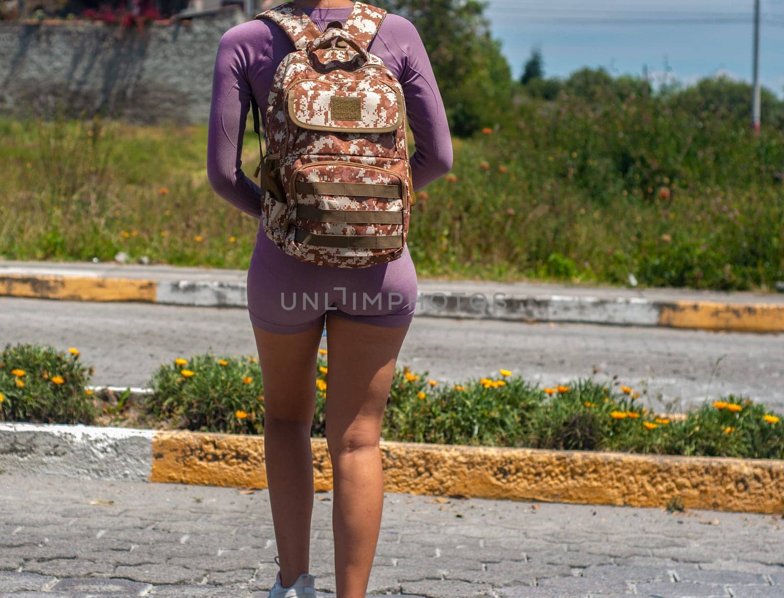 A woman wearing a purple outfit and a patterned backpack stands on the edge of an asphalt road, poised as if waiting or about to embark on a journey. by Raulmartin