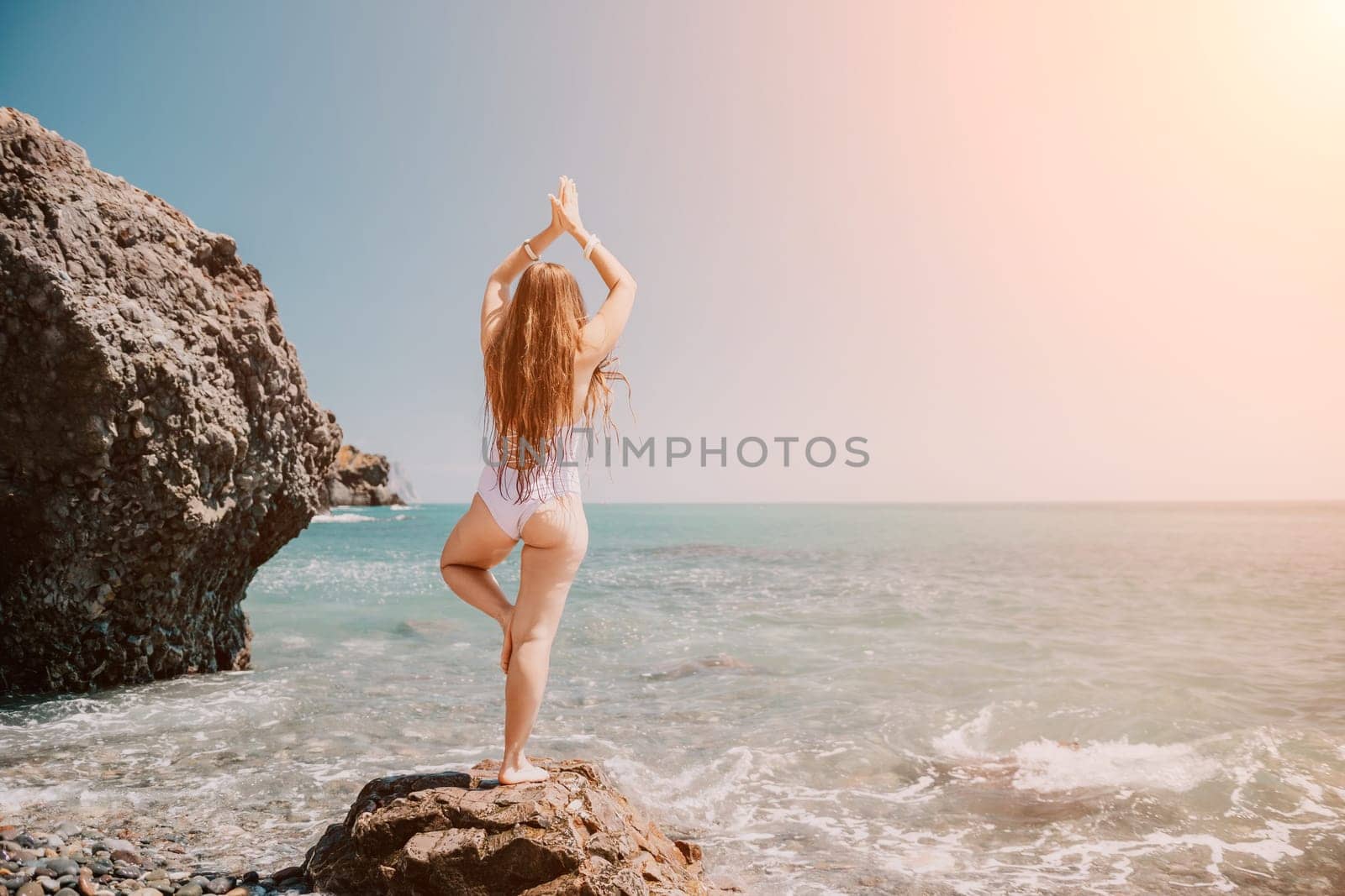 Woman sea yoga. Back view of free calm happy satisfied woman with long hair standing on top rock with yoga position against of sky by the sea. Healthy lifestyle outdoors in nature, fitness concept. by panophotograph