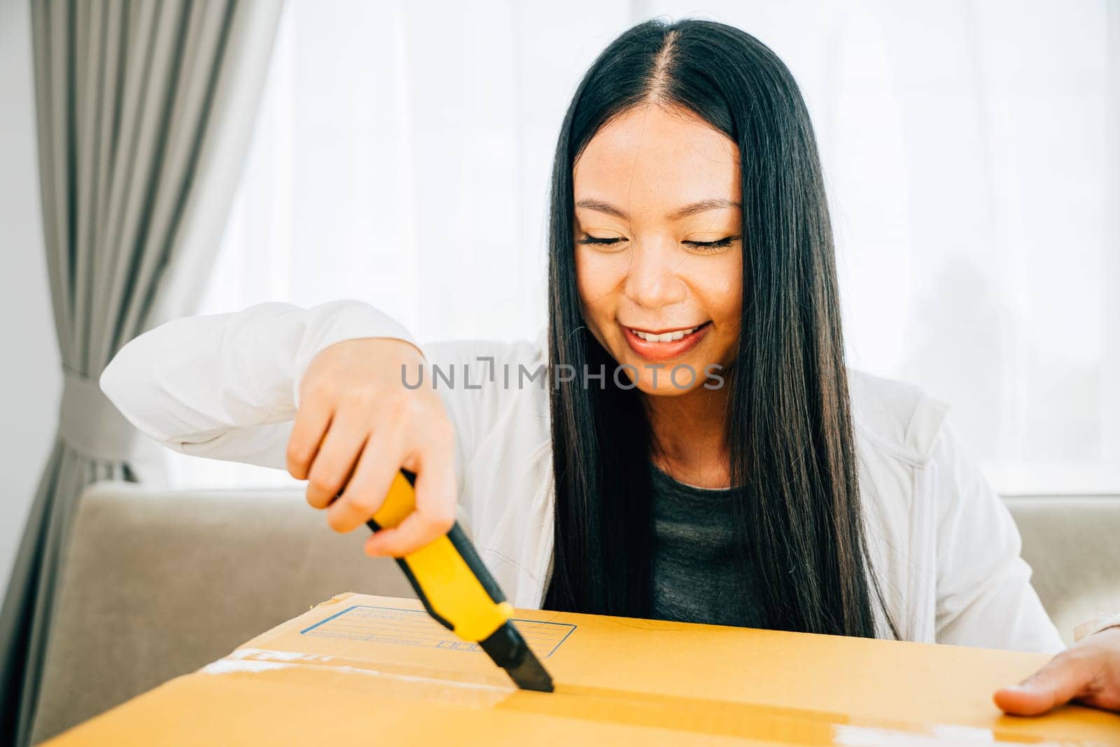 A woman on a sofa uses a cutter for precision unboxing revealing online shopping contents. Engaged in opening unpacking and discovering. Retail and surprise concept.