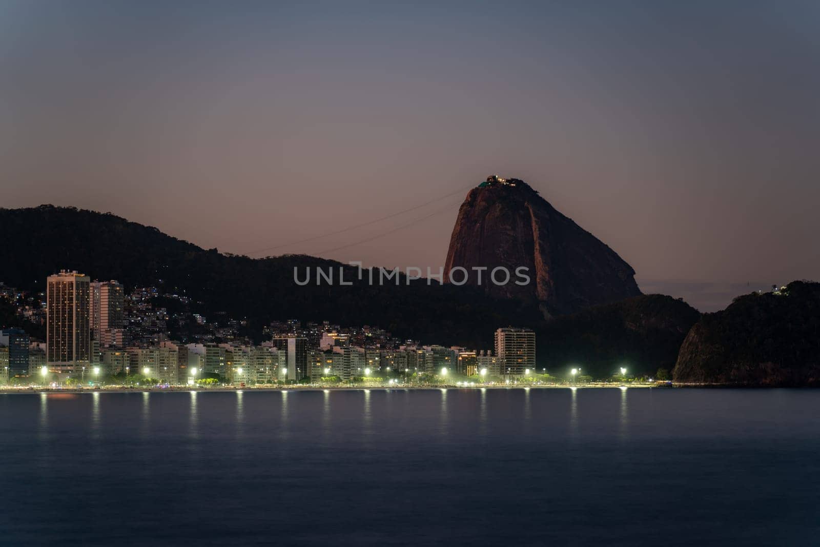 Evening falls on a city, highlighting Sugarloaf Mountain and a lit-up seaside.