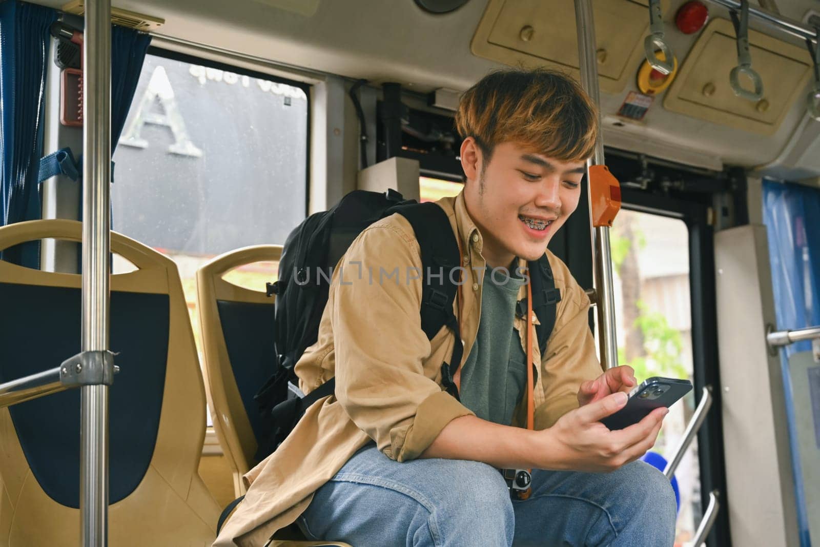 Happy young Asian man with backpack commuting by public transport and using mobile phone.