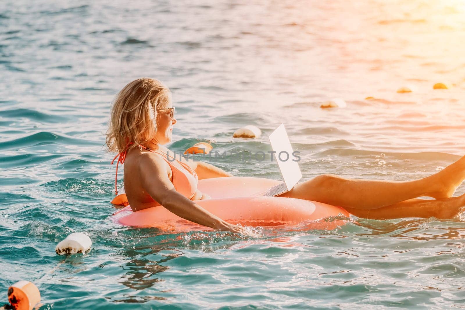 Woman works on laptop in sea. Freelancer, young blond woman in sunglases floating on an inflatable big pink donut with a laptop in the sea at sunset. Freelance, travel and holidays concept