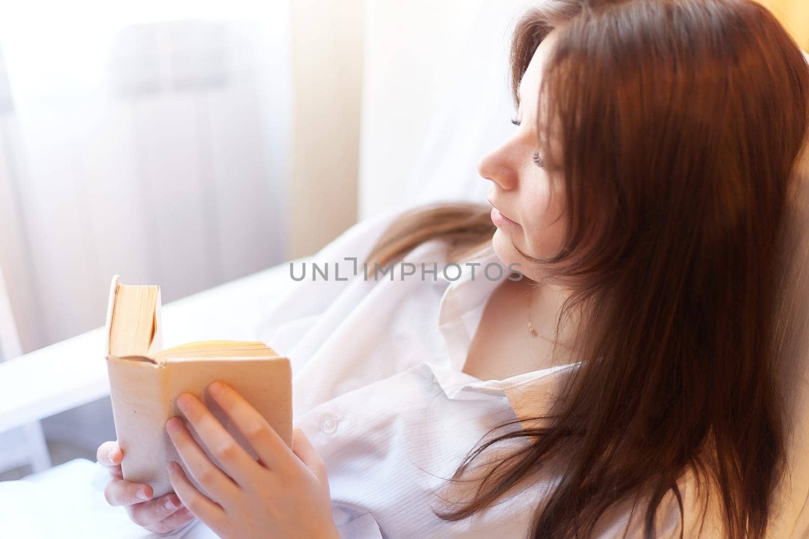 A cute brunette girl is reading a small book of poetry in the bedroom in the morning. A young pregnant woman in the maternity hospital with a book of tips on parenting. A sick and recovering lady