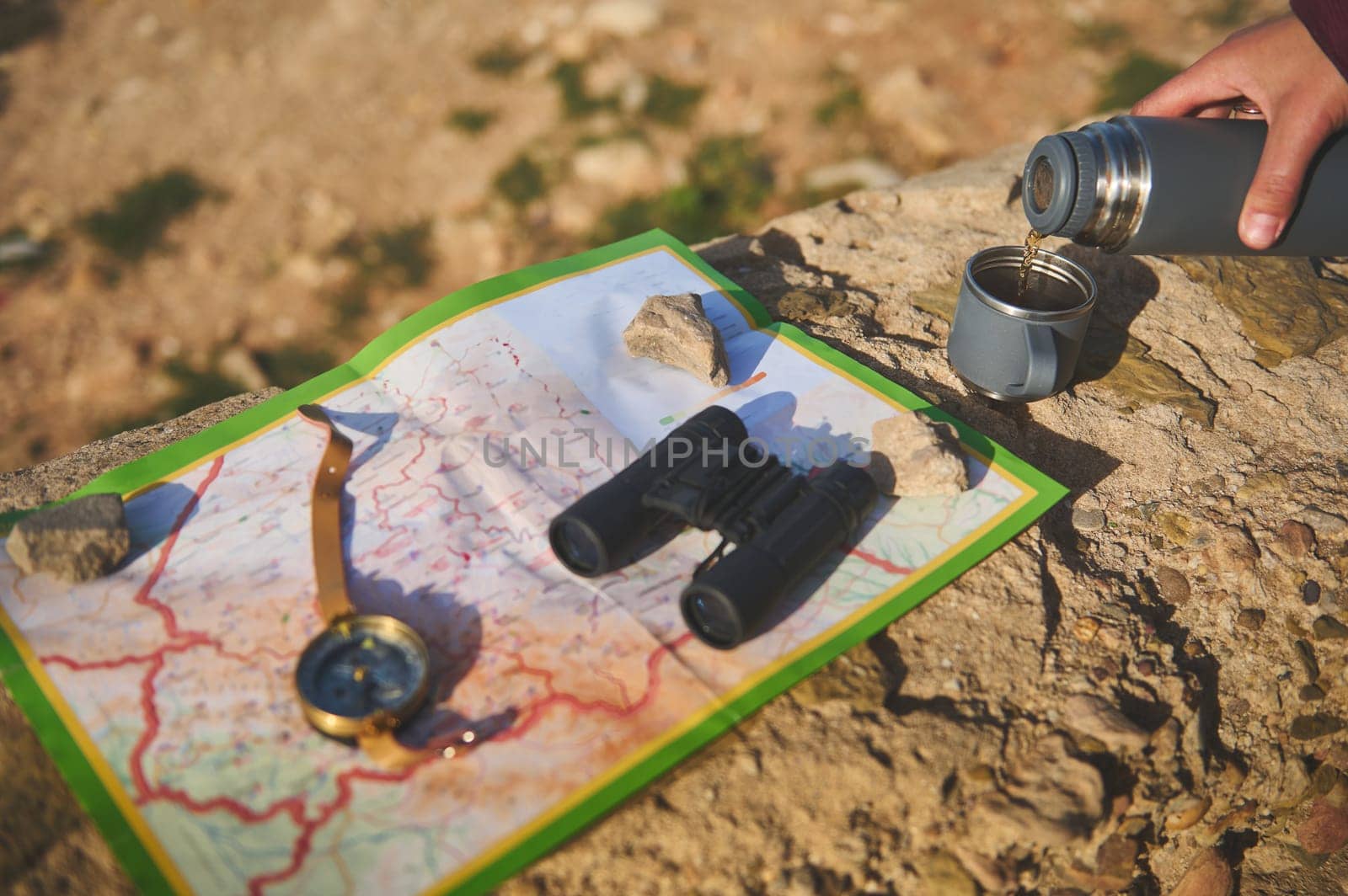 Details on tourist hand holding thermos flask and pouring hot tea drink into a stainless steel mag, standing on a rock near a map, compass and binoculars. Active lifestyle. Recreation. Tourism concept