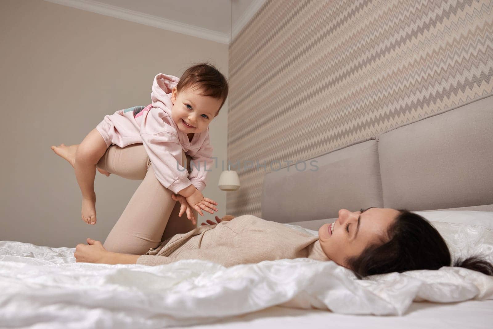 Happy Caucasian mother raising laughing cute baby and playing together in bedroom