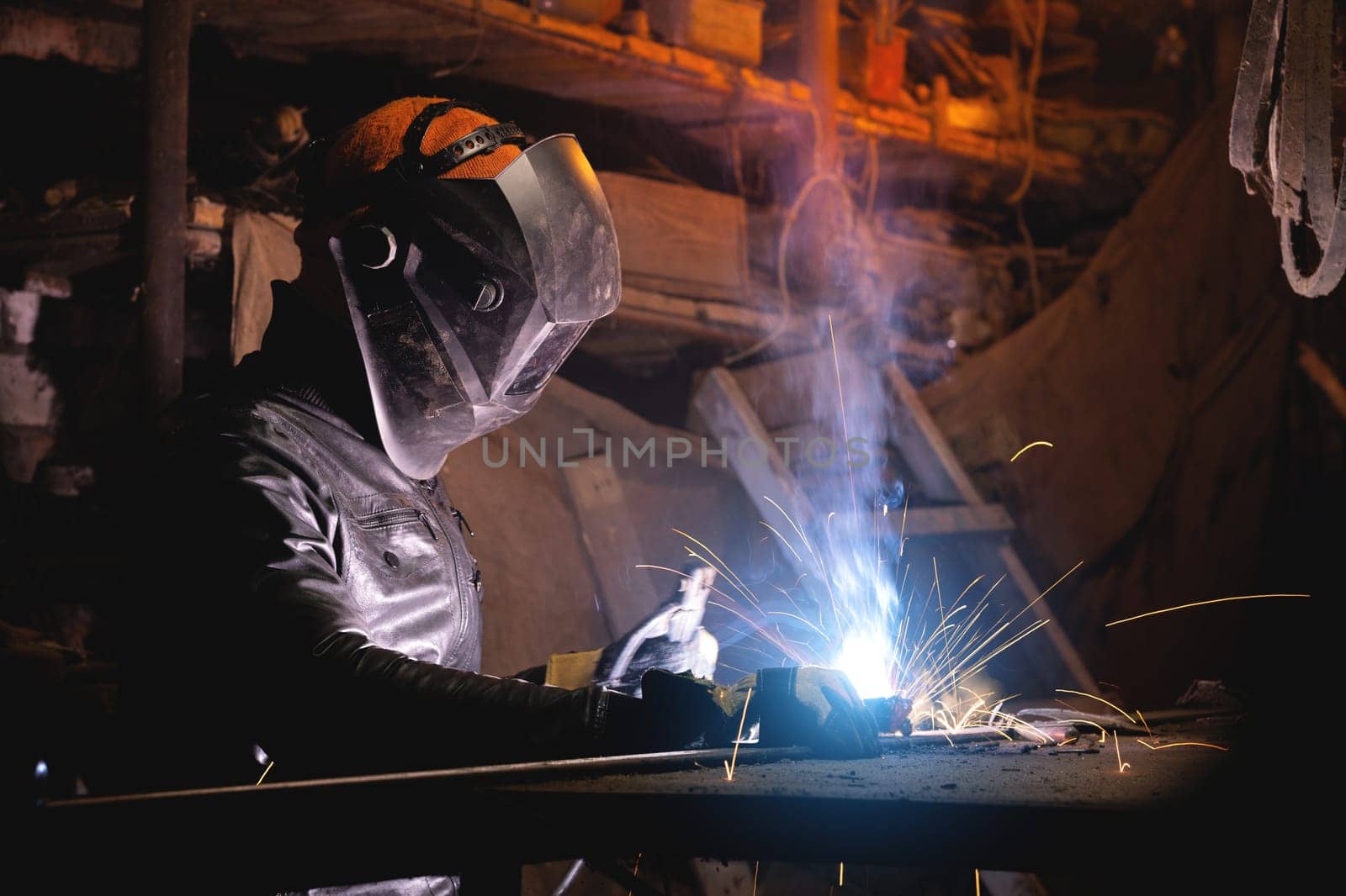 A male welder in a leather jacket carries out welding work in a home shed against the background of a brick wall. Handicraft production of metal structures and products.