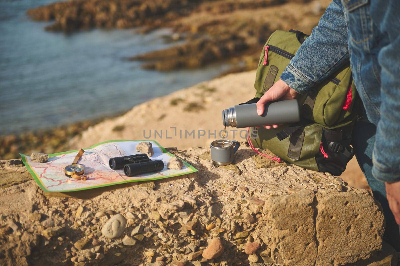 Close-up tourist hand holding thermos flask and pouring hot tea drink into a stainless steel mag, standing on a rock near a map, compass and binoculars. Active lifestyle. Recreation. Tourism concept