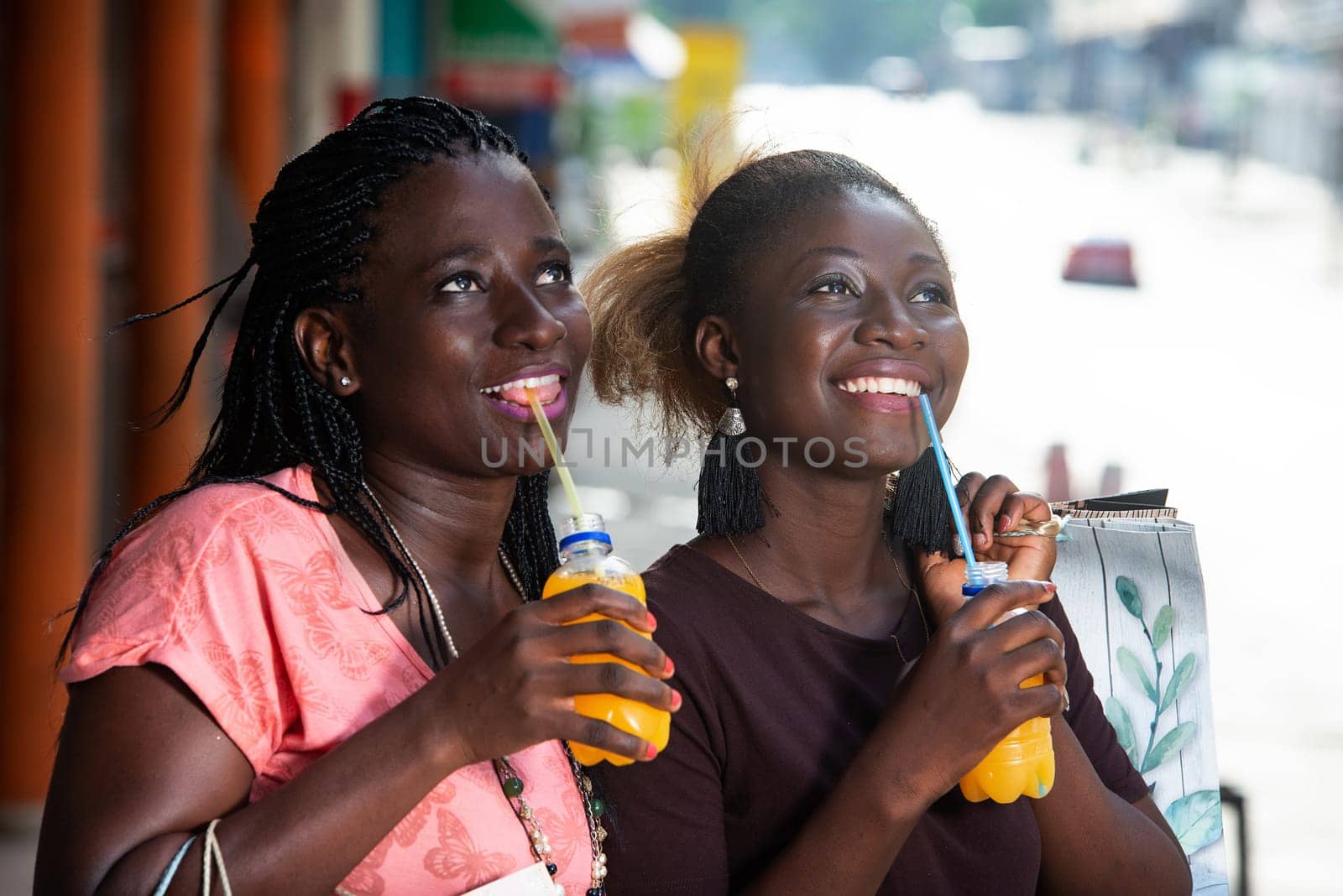 Young girls standing outdoors looking at something up there smiling with shopping bags and bottles of juice in hand.