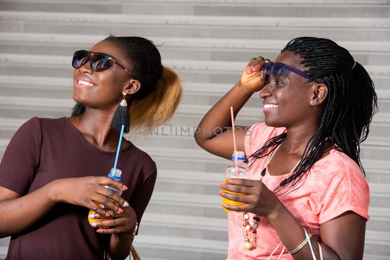 Young girls standing in sunglasses with bottles of fruit juice watching something with shopping bags in hand while smiling.