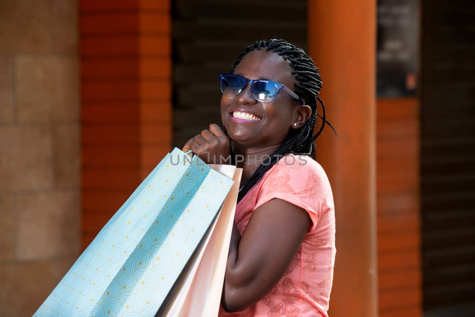 young woman standing outdoors in sunglasses laughing with shopping bags in hand.