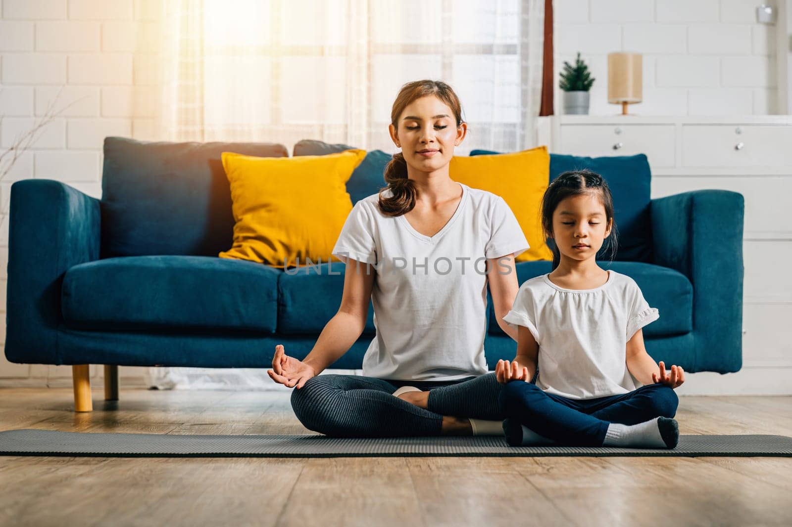 A beautiful young woman and her charming daughter share smiles during their family yoga session at home emphasizing mindfulness and meditation in lotus position creating happiness and togetherness.