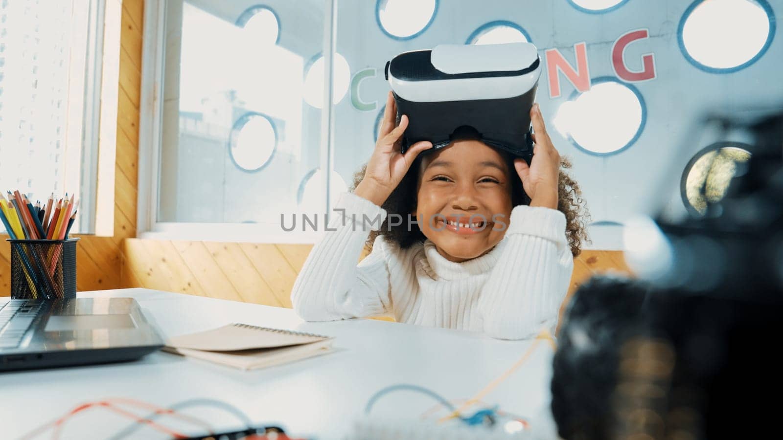African girl taking off VR glass or head set while looking at camera. Student smiling at camera with colored pencil and laptop placed on table in STEM technology class. Online education. Erudition.