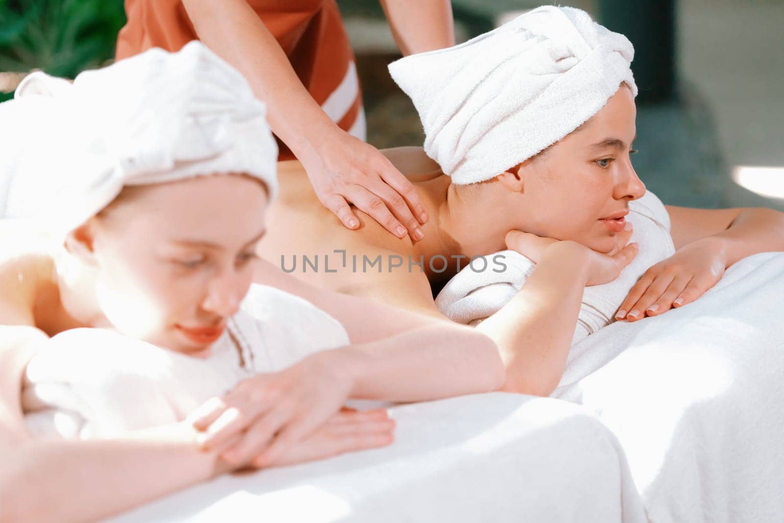 A couple of beautiful young woman with white towel lie on comfortable spa bed during receive relaxing body massage by professional masseur surrounded at spa salon. Gray background. Tranquility.