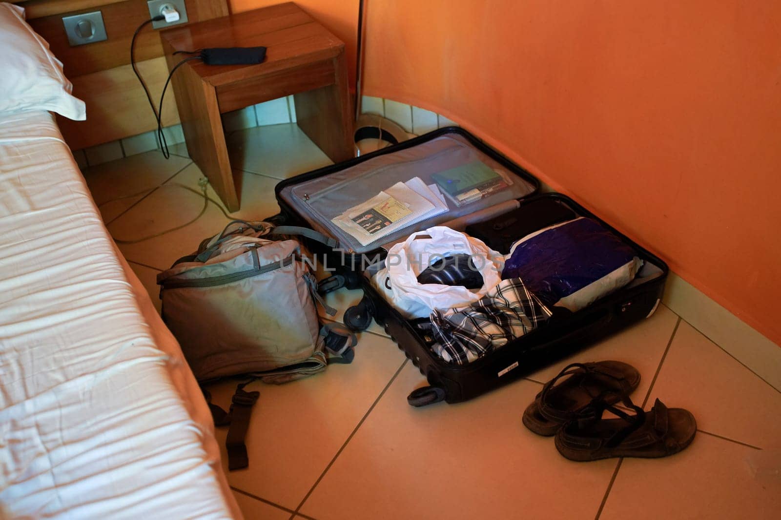 Untidy black luggage suitcase opened on hotel room floor, backpack and pair of sandals next to it.