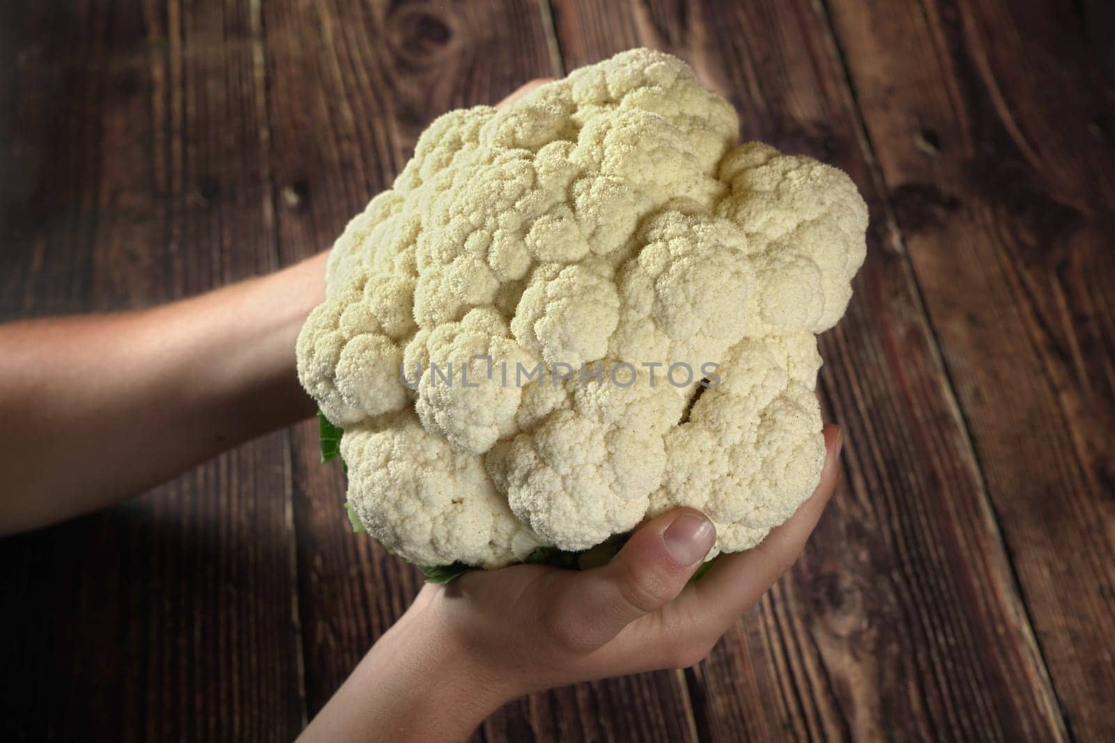 Hands holding cauliflower head over dark wooden board by Ivanko