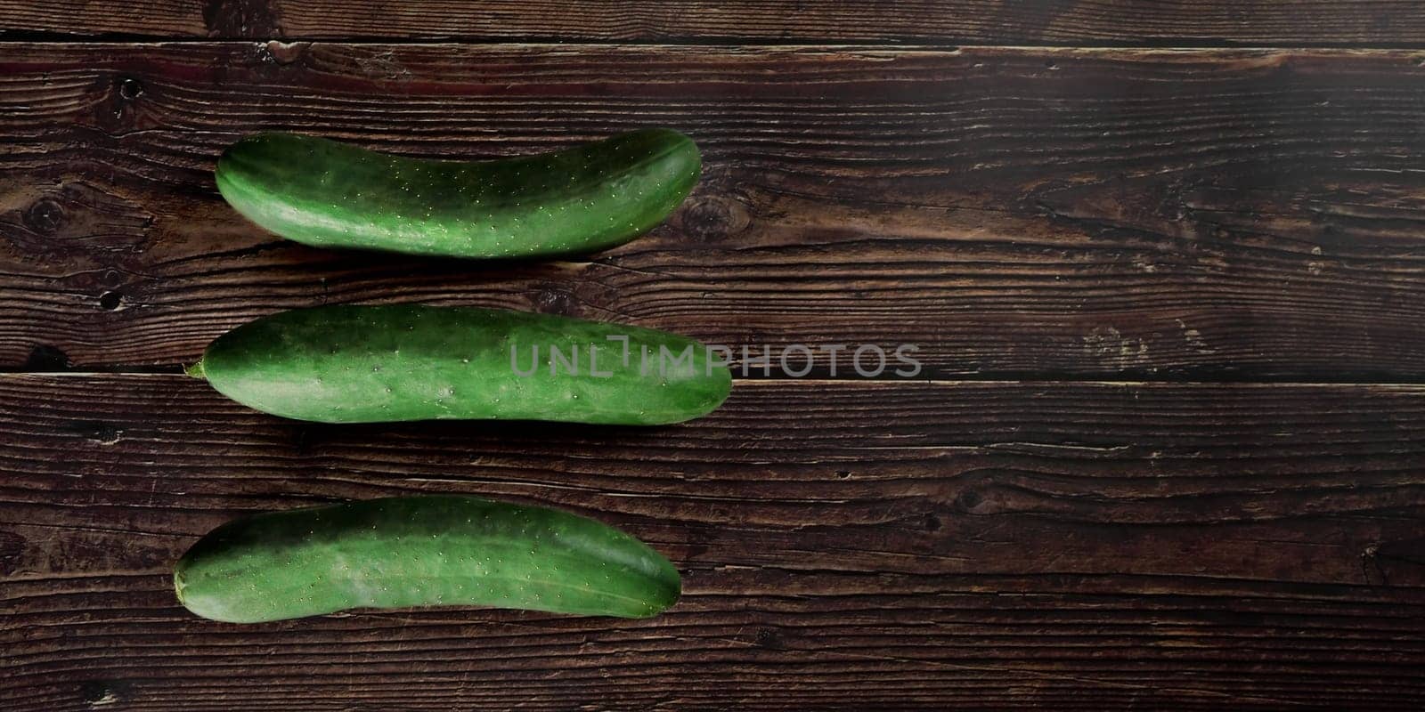 Three green cucumbers laying on dark wooden table, view from above, wide banner, space for text right side