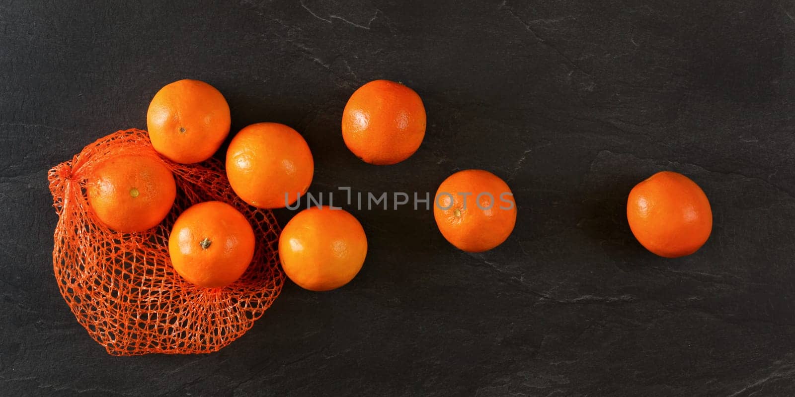 Top down view - oranges in net, some scattered on black stone like board by Ivanko