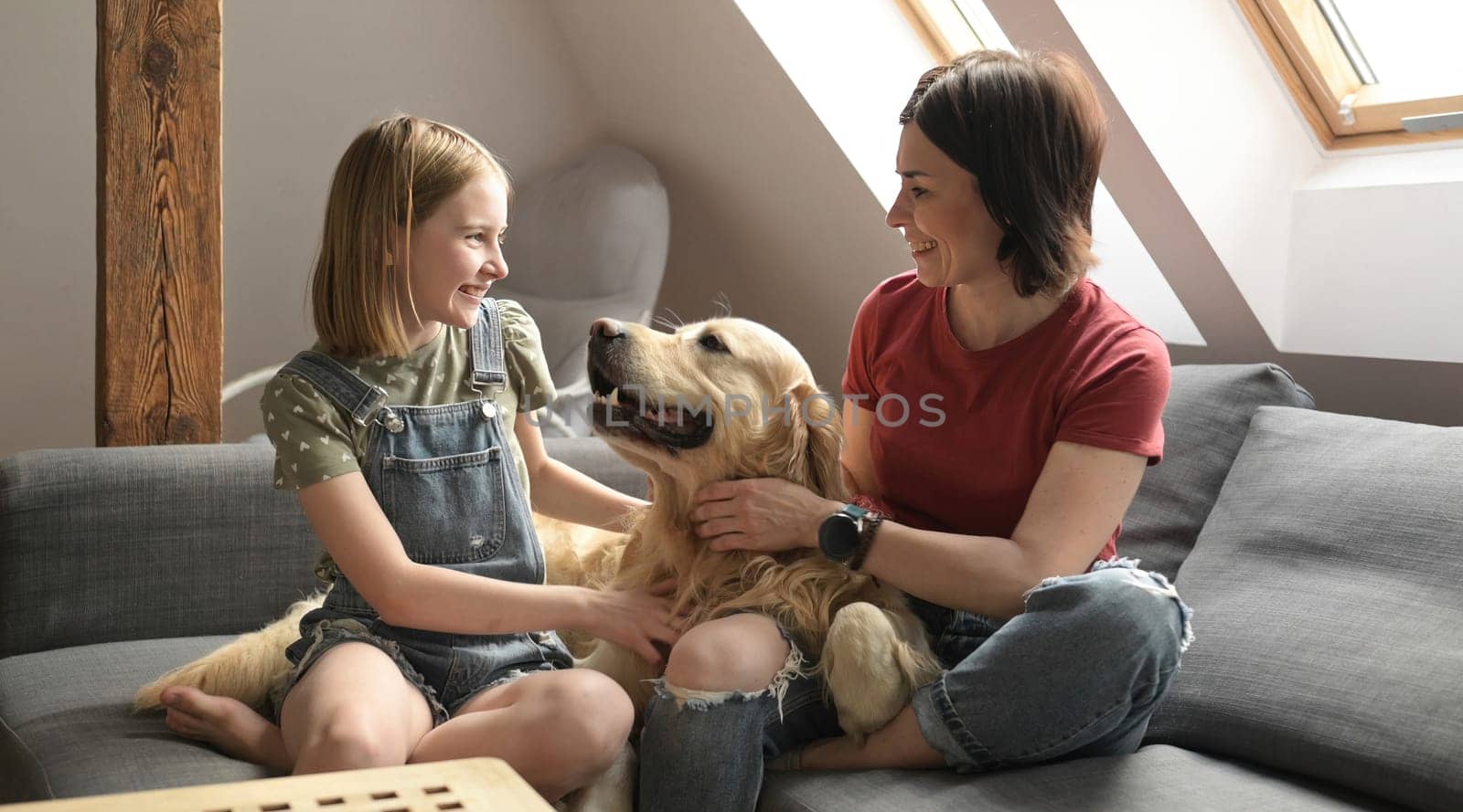 Pretty girl and preteen child family petting golden retriever dog sitting on sofa. Mother and daughter with purebred pet doggy at home smiling