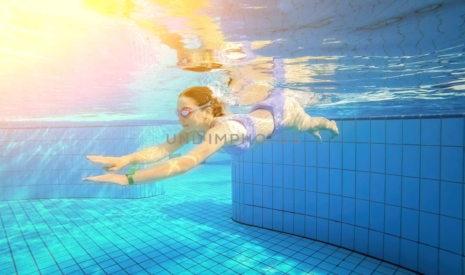 Beautiful girl swimming under water in blue pool wearing swimsuit and googles. Pretty teenager enjoying diving lessons