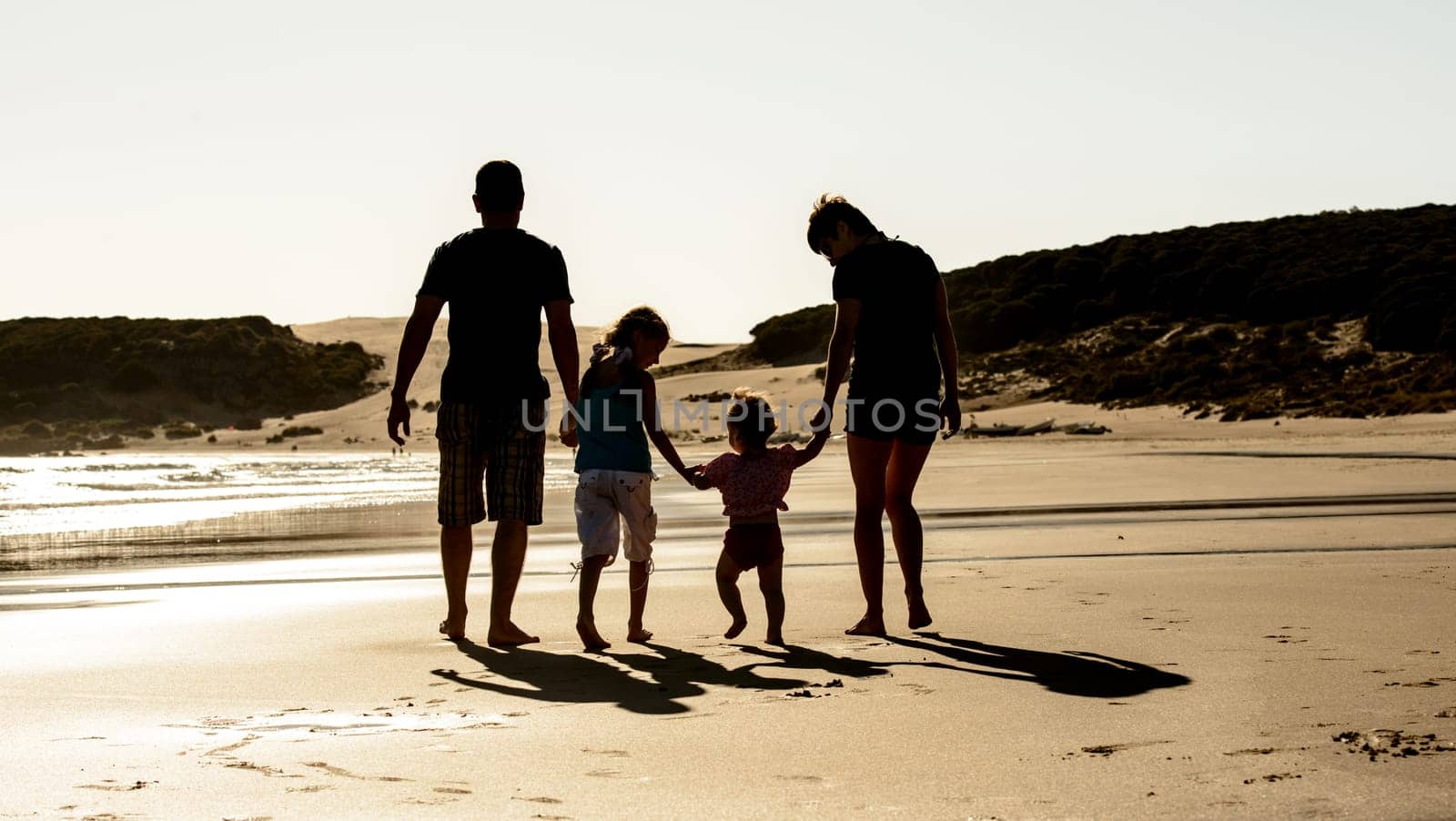 Happy family with children silhouette at sea beach sunset. Father, mother and kids walking on seashore with evening sun light