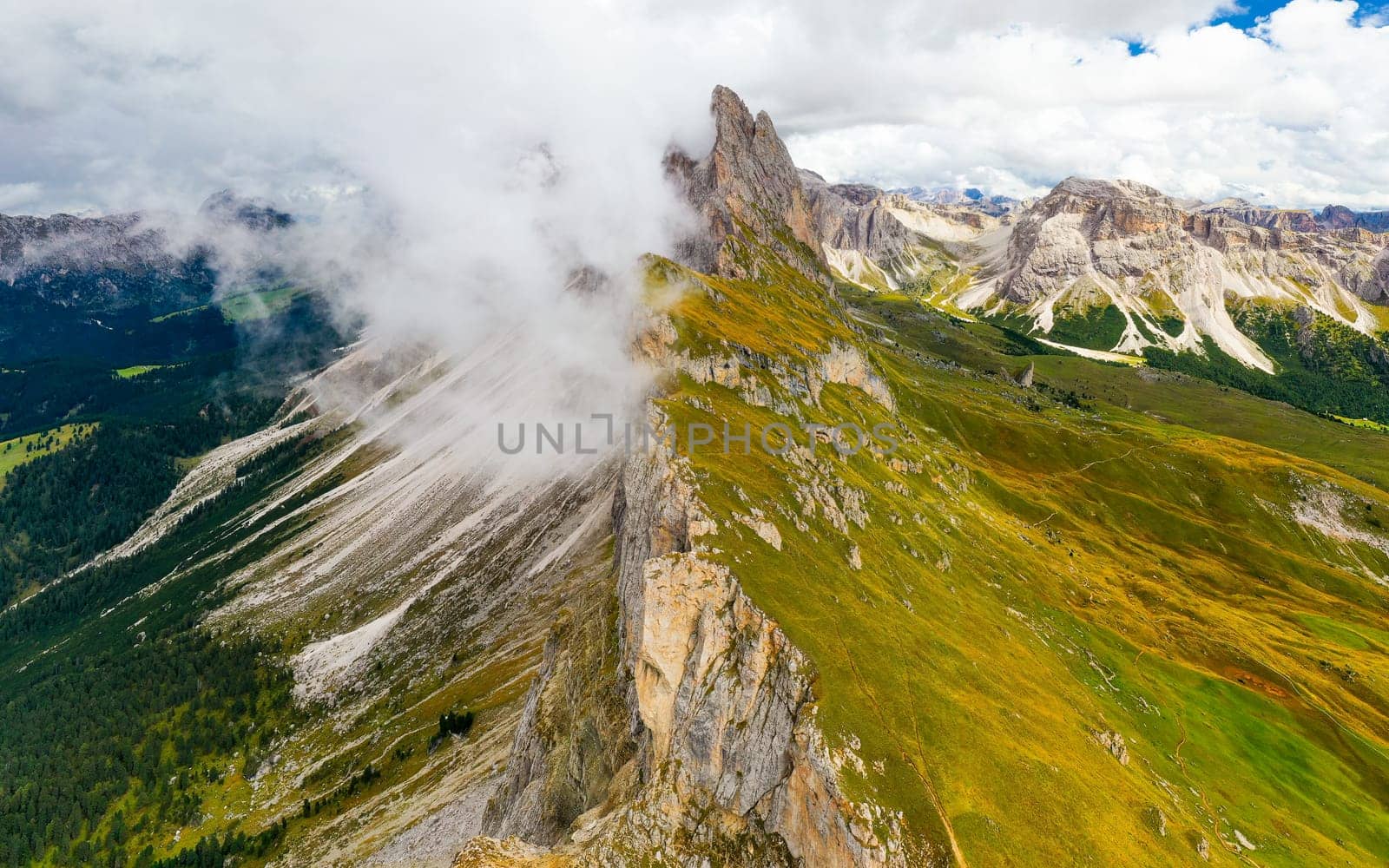 Dense foggy cloud descends on giant Seceda ridgeline with green slopes and bare peak. Cloudy sky above touristic Italian Alps aerial view