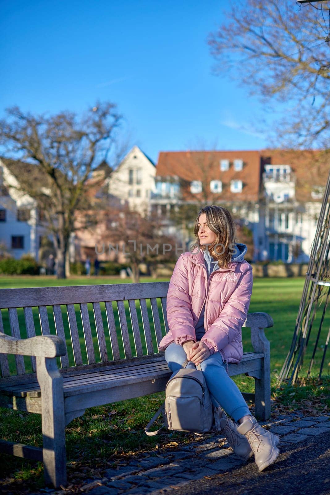 Winter Joy in Bitigheim-Bissingen: Beautiful Girl in Pink Jacket Sitting Amidst Half-Timbered Charm. beautiful girl in a pink winter jacket sitting on a bench in a park, set against the backdrop of the historic town of Bitigheim-Bissingen, Baden-Württemberg, Germany. by Andrii_Ko