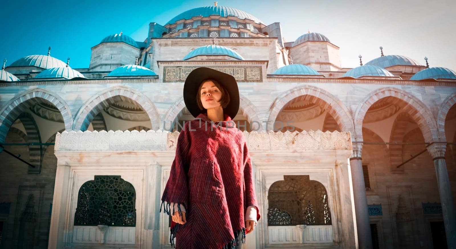 A young girl traveler in a stylish red poncho and a hat stands against the background of a blue mosque in Istanbul, Turkey. Backpacker travels to Arab countries