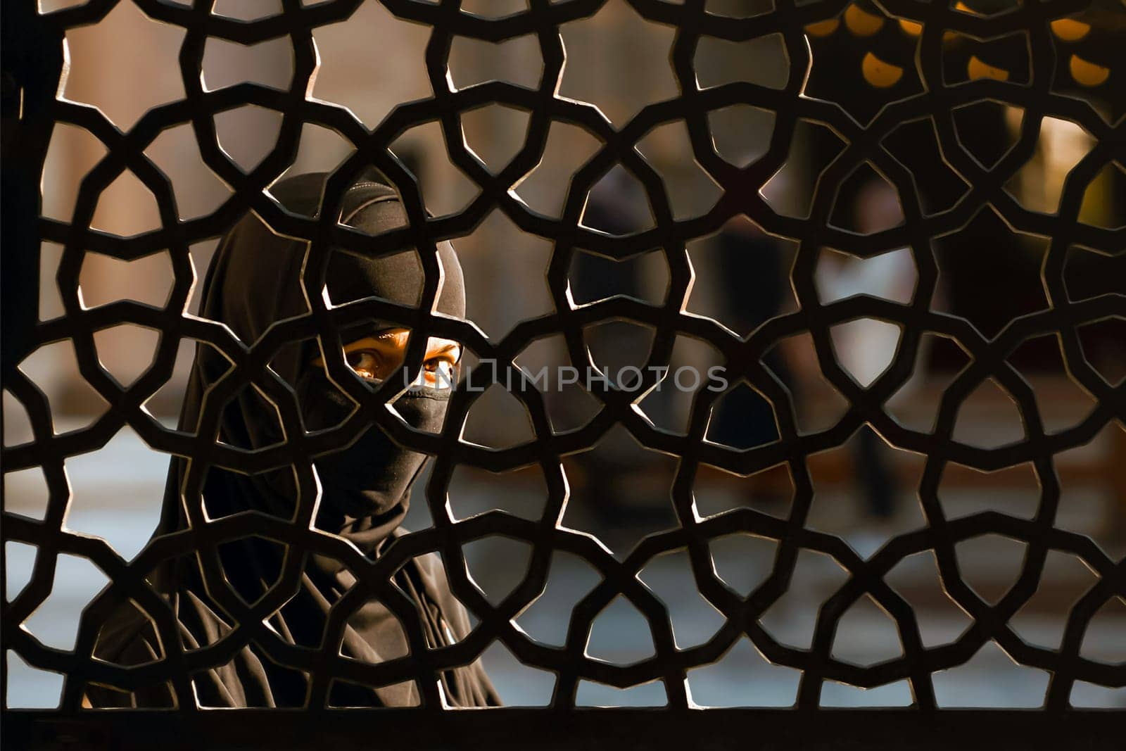 A mysterious girl in a hijab stands behind an ornate wrought-iron fence in a religious complex near with mosque. Travel to Eastern countries. Turkey