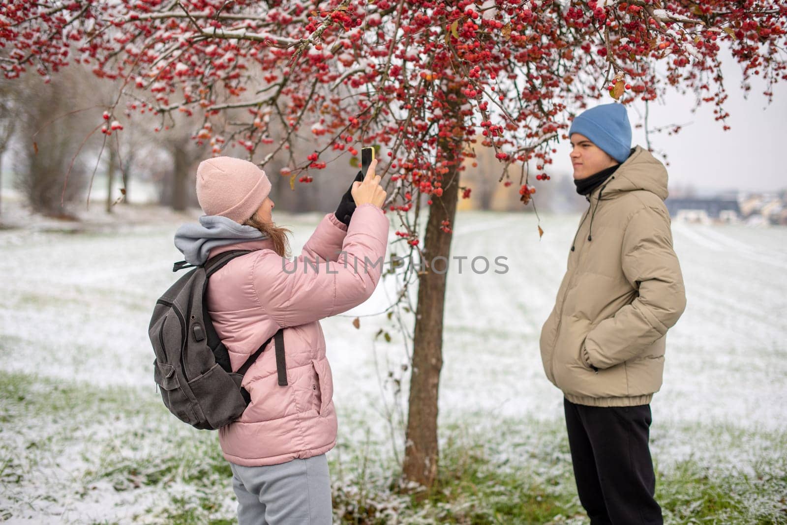 Winter Romance: Girl in Pink Winter Jacket Photographing Boy Against Snow-Covered Red Tree and Field. Embrace the winter magic in this enchanting image, where a girl in a pink winter jacket captures a moment as she photographs her companion against the backdrop of a snow-covered red tree and field. The photograph beautifully conveys the essence of winter romance and the serene beauty of a snowy landscape.