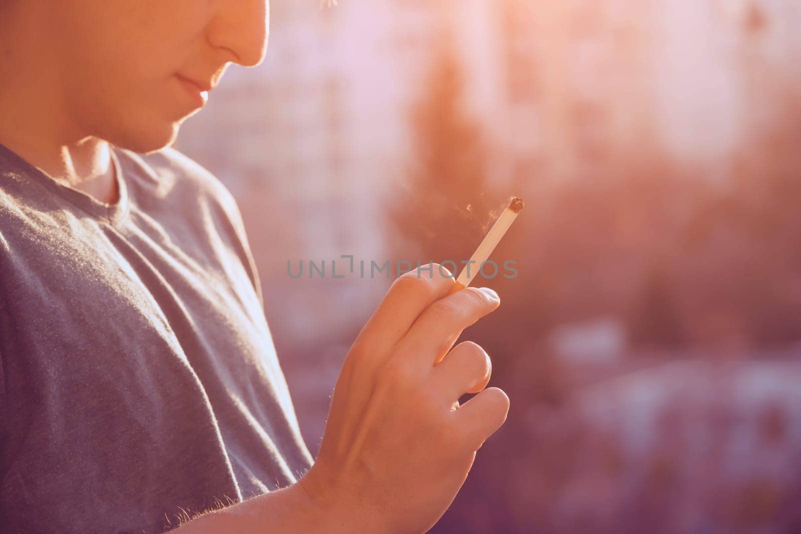 A young man smokes, holds a cigarette in his hand and inhales the smoke while standing at the window early in the morning in the sun, close-up view.