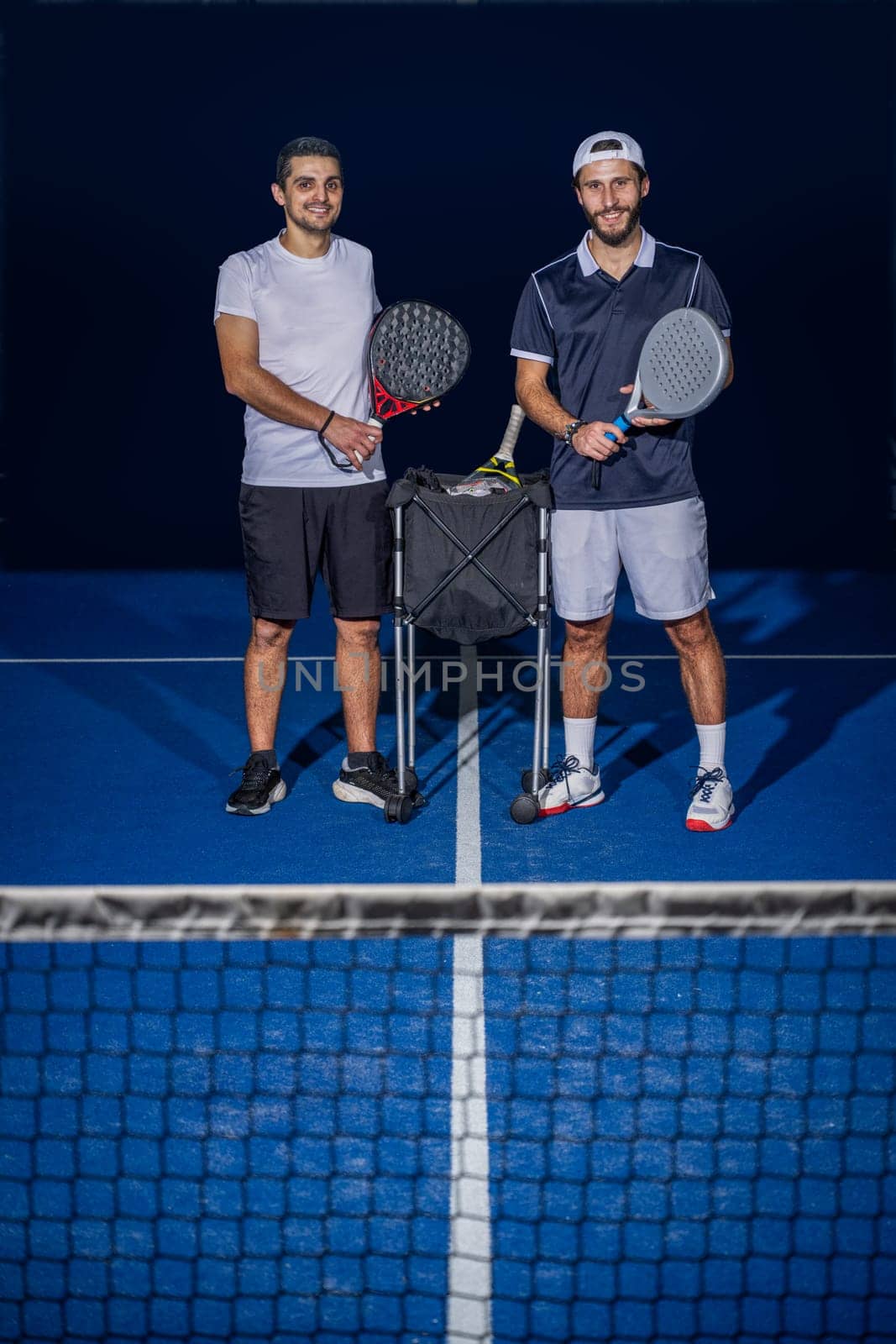 Full length portrait of two sporty young men in sportswear standing on indoor court with paddle rackets in hands after match.