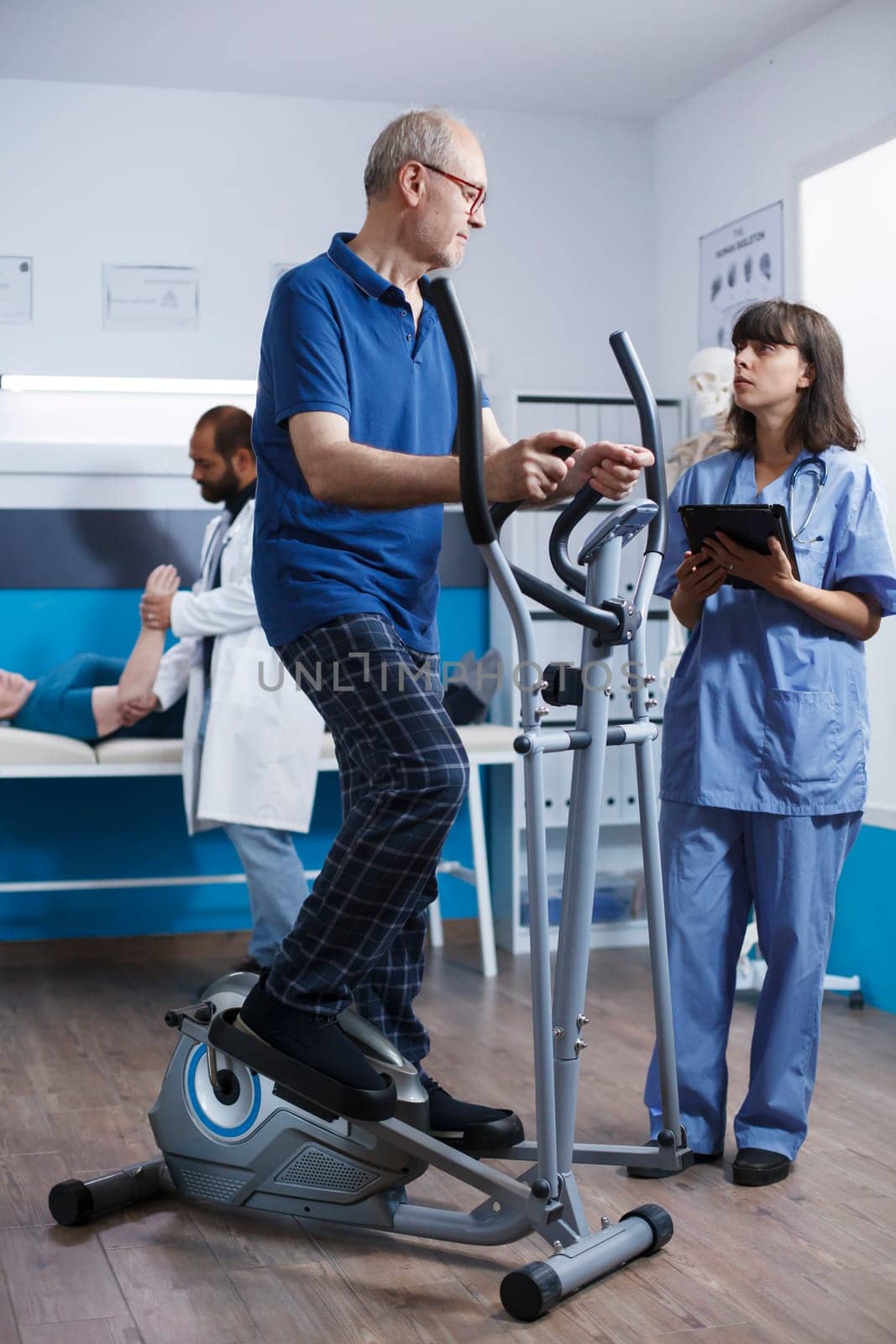 Elderly male patient undergoing physiotherapy on stationary bicycle, with assistance of nurse in blue scrubs. Medical assistant using electronic tablet assists senior citizen with physical therapy.