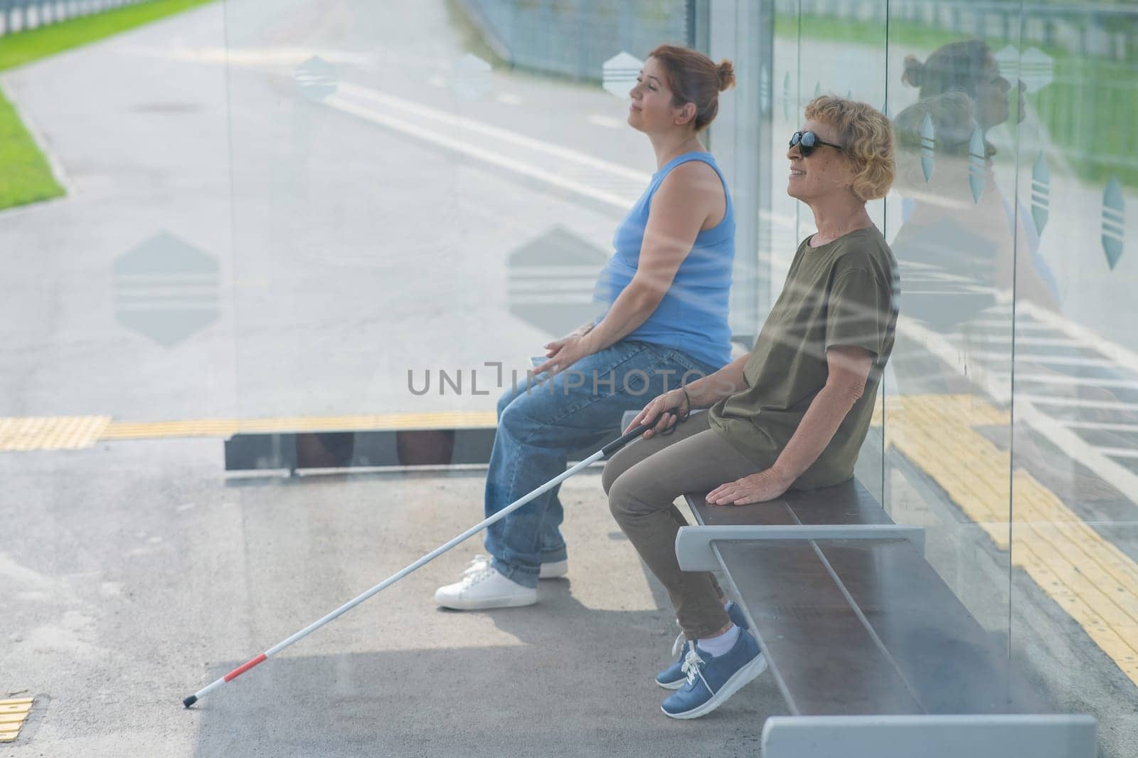 An elderly blind woman and a mature pregnant woman are sitting at a bus stop and waiting for the bus