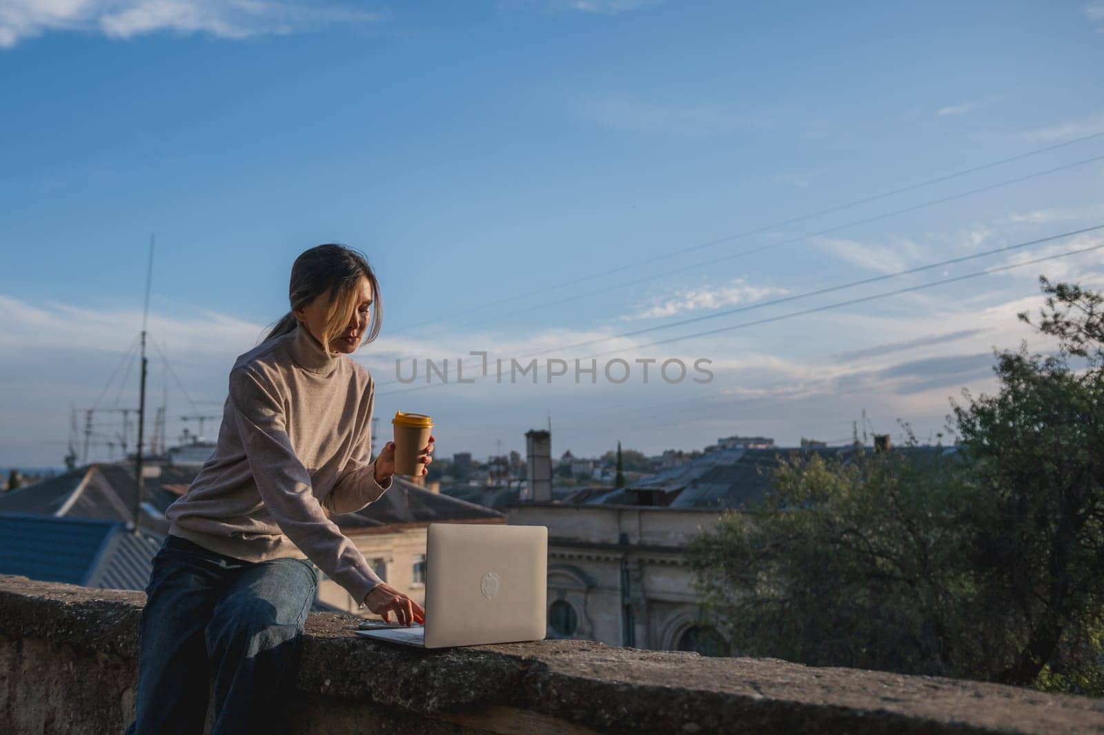 Woman freelancer uses laptop on cement wall outdoors against the sky and the roof of the city. The woman to be focused on her work or enjoying some leisure time while using her laptop