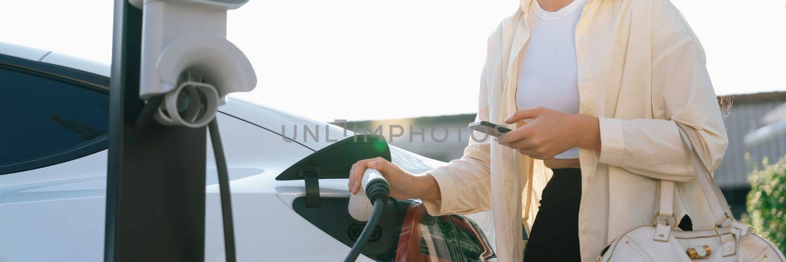 Woman holding shopping bag and use smartphone to pay for electricity to recharge EV car battery from charging station at city mall parking lot. Modern woman go shopping by eco car.Panorama Expedient