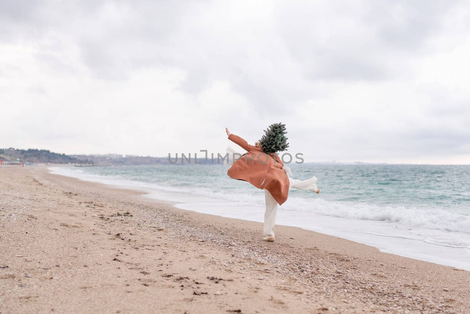 Blond woman Christmas tree sea. Christmas portrait of a happy woman walking along the beach and holding a Christmas tree on her shoulder. She is wearing a brown coat and a white suit. by Matiunina