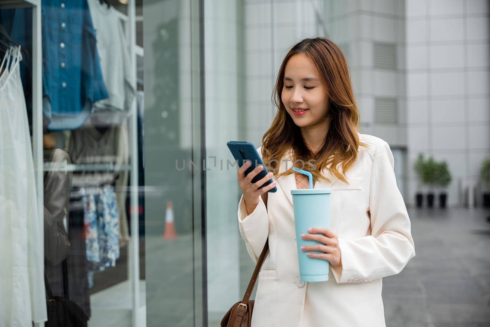 Asian woman enjoying her coffee and digital communication ,holding a tumbler mug and smartphone in front of a store window. Modern lifestyle and convenience is her style.