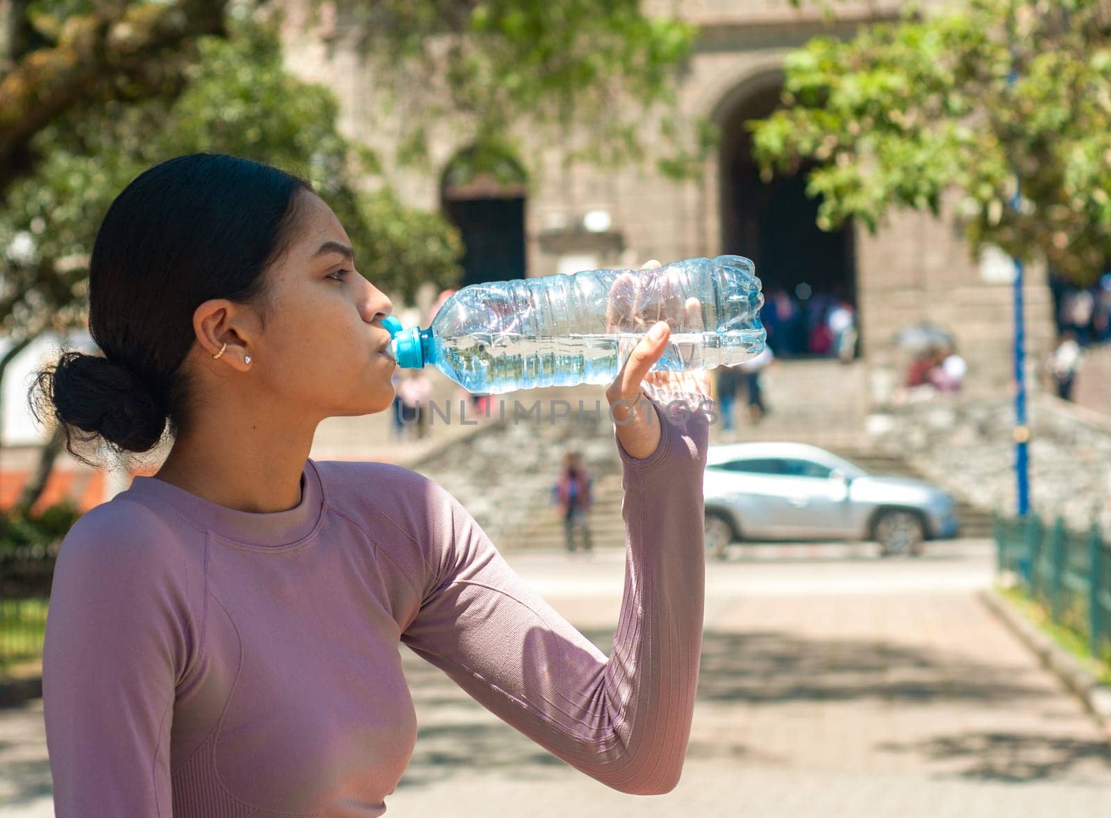 A Latina woman elegantly drinks cool, clear water from a plastic bottle in the city center during a quiet moment in her solo tourist experience. by Raulmartin