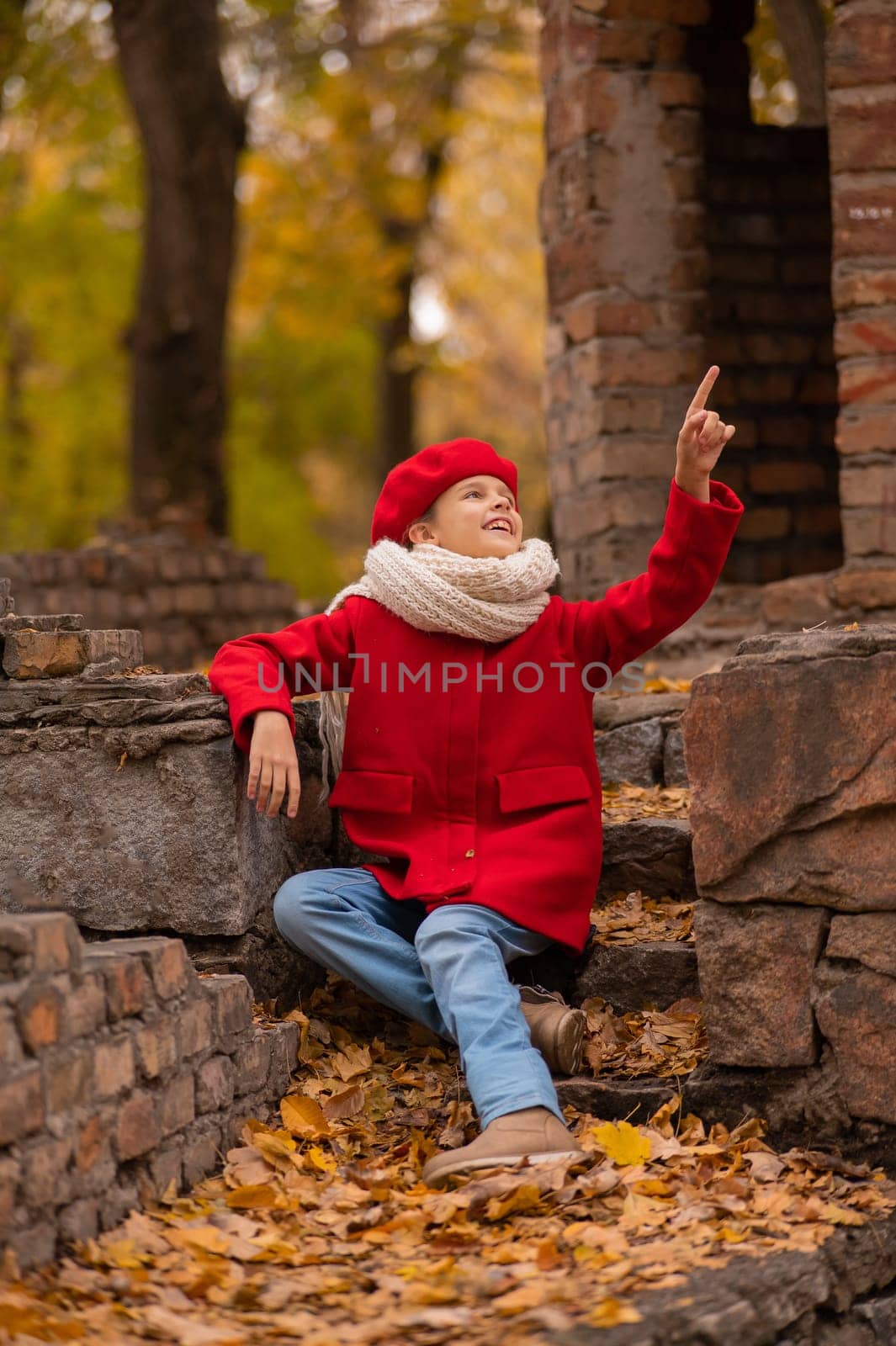 Smiling caucasian girl in a red coat and beret sits on a brick wall on a walk in autumn