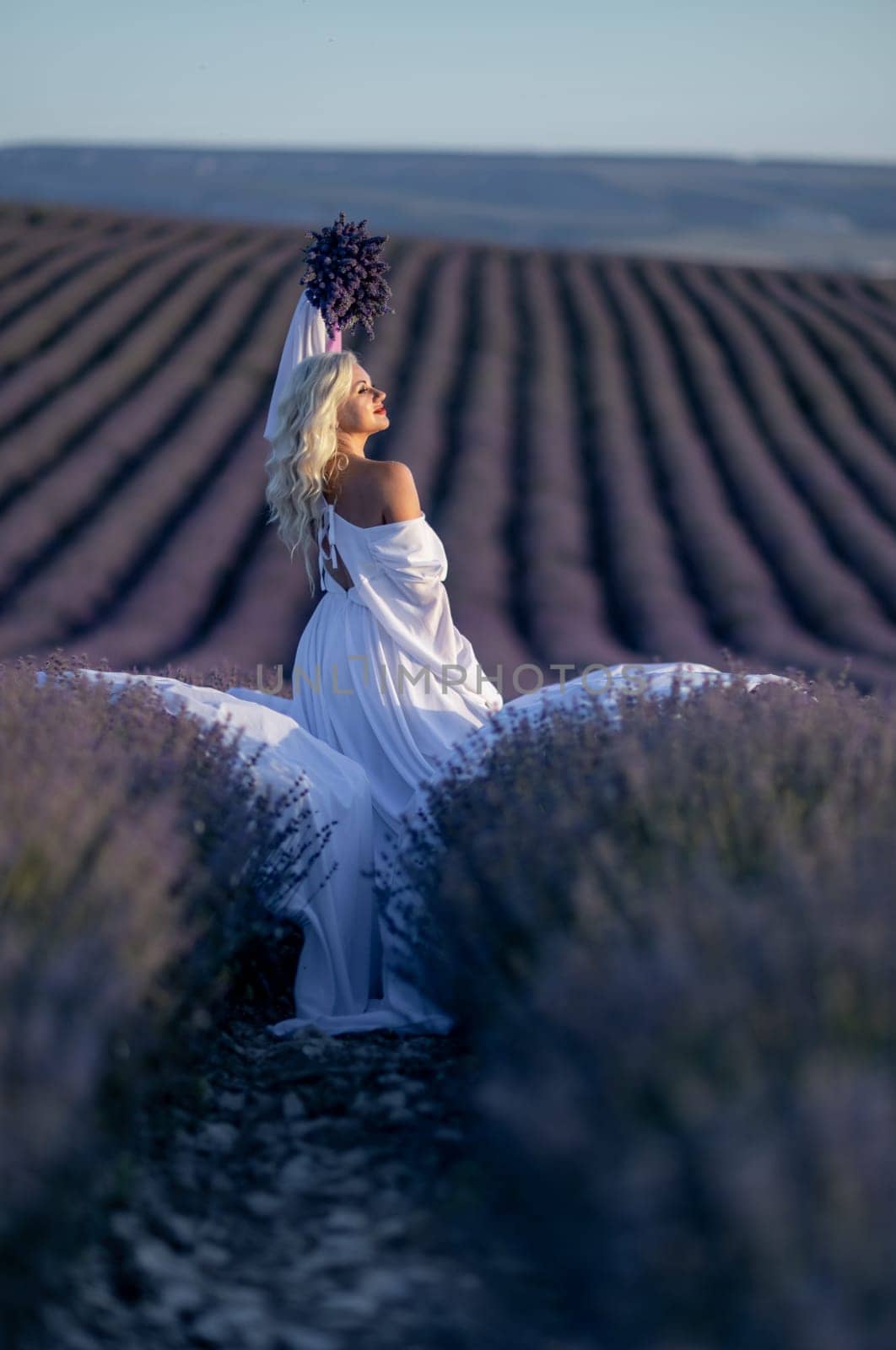 Blonde woman poses in lavender field at sunset. Happy woman in white dress holds lavender bouquet. Aromatherapy concept, lavender oil, photo session in lavender by Matiunina